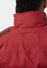 Czerwona wodoodporna kurtka z wysokim kołnierzem, charakteryzująca się subtelna teksturą i czarną marką na lewym ramieniu. Szczegóły obejmują tylnią kieszeń.