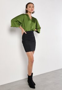 Grünes Satinbluse mit Knopfverschluss, weiten Ärmeln; schwarze geraffte Minirock. Schwarze Stiefeletten runden den Look auf einem hellgrauen Hintergrund ab.