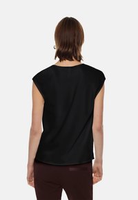 Top nero a maniche corte con texture liscia e scollatura rotonda, caratterizzato da una vestibilità rilassata e un orlo leggermente curvo. Indossato con pantaloni bordeaux scuri.