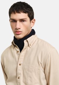 Camisa de botones beige con un bolsillo en el pecho, combinada con un suéter de cuello alto oscuro. La tela parece suave y tiene una textura sutil.