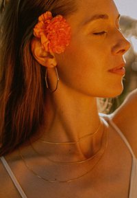 Goldene, schichtweise angeordnete Halsketten, ein achteckiger Ohrhänger und eine orange Blume betonen das sonnenbeschienene Profil einer Frau mit geschlossenen Augen.