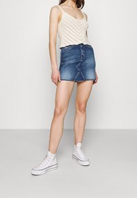 Ljust beige stickad tanktopp med v-ringning och zigzagmönster, tillsammans med en mid-blå denim-minikjol med rå fåll och sömdetalj fram. Vita högklackade sneakers.