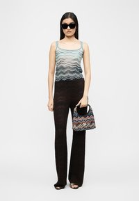 Γυναίκα με εμπριμέ αμάνικο τοπ και μαύρο παντελόνι κρατά πολύχρωμη τσάντα με ζικζακ σχέδιο, φοράει μαύρα γυαλιά ηλίου και σανδάλια.