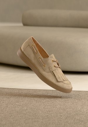 Beige suède loafer met franjes en kanten details die zweeft boven een bijpassend gestructureerd tapijt in een neutraal getinte binnenruimte.