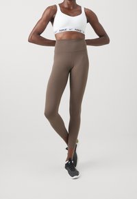 Bruna höga leggings i ett mjukt, stretchigt material, kombinerat med en vit sport-bh med ett svart logoband.