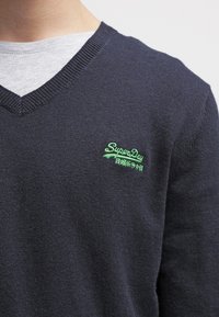 Granatowy sweter w serek wykonany z dzianiny, z małym zielonym haftowanym logo po lewej stronie klatki piersiowej, noszony na szarym T-shircie.