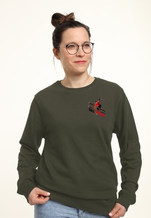 Kobieta w okularach i z włosami splecionymi w kok nosi oliwkową bluzę z małym czerwonym narciarzem i napisem „Ski Style” na piersi, stojąc na tle jasnego tła.