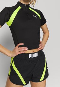 Černý sportovní top s krátkými rukávy a půlzipem, doplněný neonově zelenými bočními panely; spárovaný s černými šortkami se zelenými pruhy a logem.