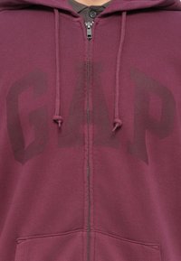 Mørkerød zip-op hættejakke med et let falmet stort "GAP" logo henover brystet og snorene på hætten.