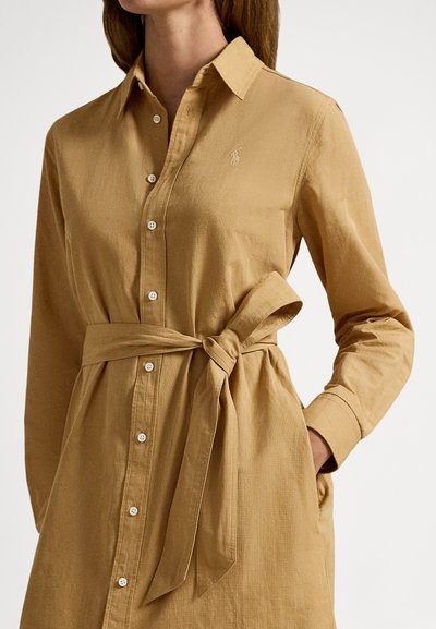 Kvinne iført en langermet kjole i kamelfarge med knapper og knytt belte rundt livet, hånden i lommen.