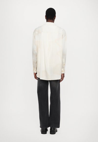 Camisa de manga larga en color crema con suaves patrones, pliegue en la espalda y puños de botones, combinada con vaqueros de pierna ancha negros y zapatos negros.