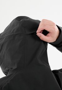 Svart vattentät jacka med huva, featuring en ren design och en hand som justerar dragsnöret. Materialet ser slätt och lättviktigt ut.