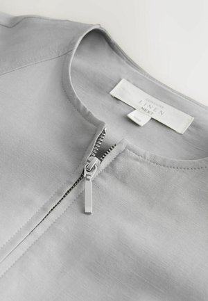 Gaiši pelēka lina krekls ar apaļu kakla izgriezumu un daļēji atvērtu sudraba metāla rāvējslēdzēju priekšpusē.