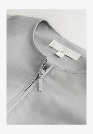 Camicia in lino grigio chiaro con scollo rotondo e cerniera metallica argentata parzialmente aperta sul davanti.