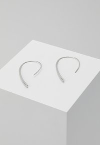 Boucles d'oreilles en argent en forme de cerceau, avec une silhouette élégante et allongée. Conçues avec une finition lisse et un design ouvert. Exposées sur un piédestal blanc.