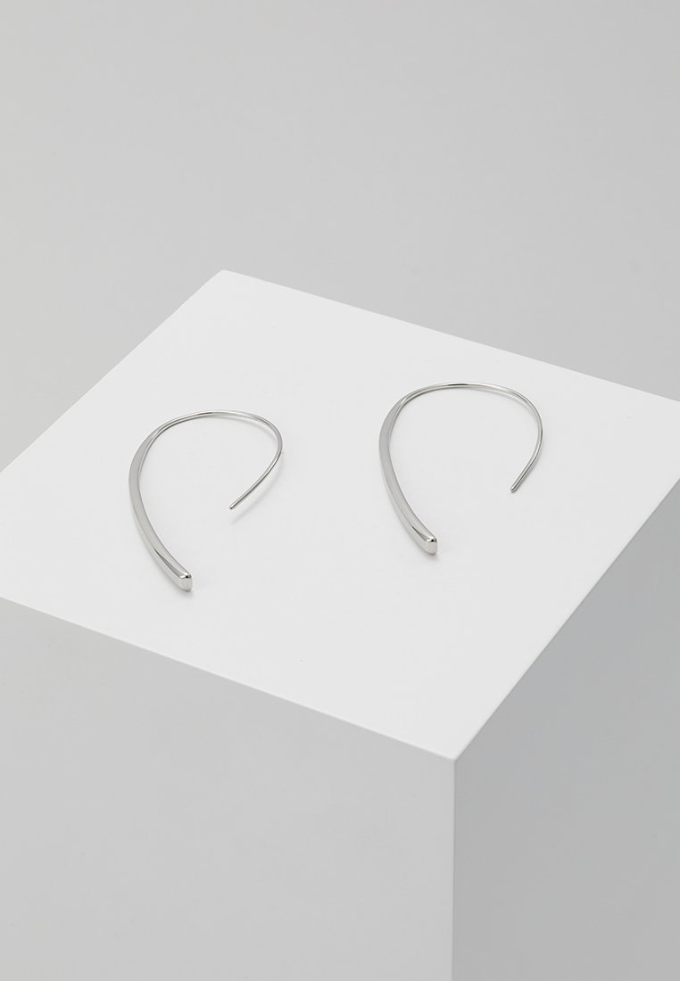 Boucles d'oreilles en argent en forme de cerceau, avec une silhouette élégante et allongée. Conçues avec une finition lisse et un design ouvert. Exposées sur un piédestal blanc.