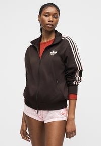 Junge Frau trägt eine schwarze Adidas-Trainingsjacke mit weißen Streifen und hellrosa Adidas-Mesh-Shorts vor einem einfarbigen Hintergrund.