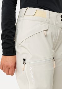 Nahaufnahme von beigen Outdoor-Hosen mit mehreren Reißverschlusstaschen und verstellbarem Klettverschluss in der Taille, getragen mit einem schwarzen Langarmshirt.