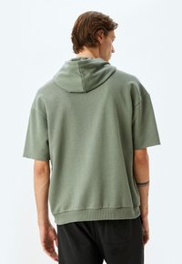 Oversized short-sleeve hoodie in olive green cotton, featuring a drawstring hood and ribbed hem. Soft texture with a relaxed fit.