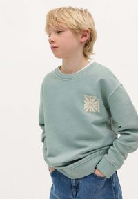 Jasnozielona bluza z ribbowanymi mankietami i dołem, z kremową grafiką na lewej piersi. Połączona z niebieskimi dżinsami.