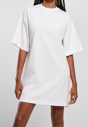 Woman wearing a plain oversized white T-shirt dress with short sleeves, standing against a plain light gray background.