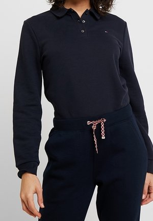 Persoon draagt een donker poloshirt met lange mouwen en een donkere trainingsbroek met een rood-wit trekkoord gestrikt in de taille.