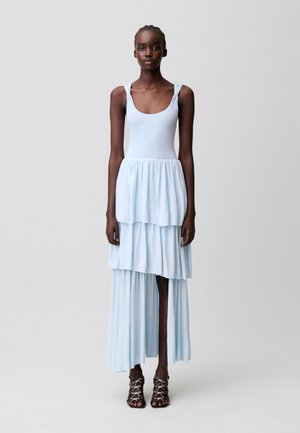 Femme grande à la peau foncée portant une robe sans manches bleu clair à volants avec un décolleté arrondi et des talons ouverts noirs à lacets.