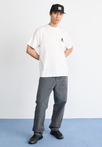 Hombre de pie con las manos detrás de la espalda, vistiendo una camiseta gráfica blanca, pantalones grises, zapatillas negras y una gorra negra, frente a un fondo blanco liso.
