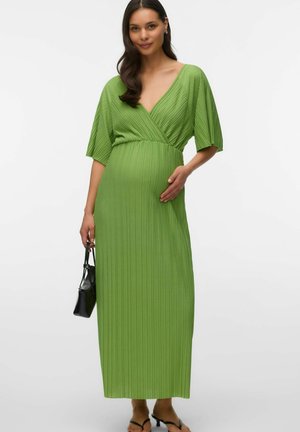 Femme enceinte vêtue d'une robe maxi plissée verte avec un col en V, tenant un petit sac à main noir et posant l'autre main sur son ventre.