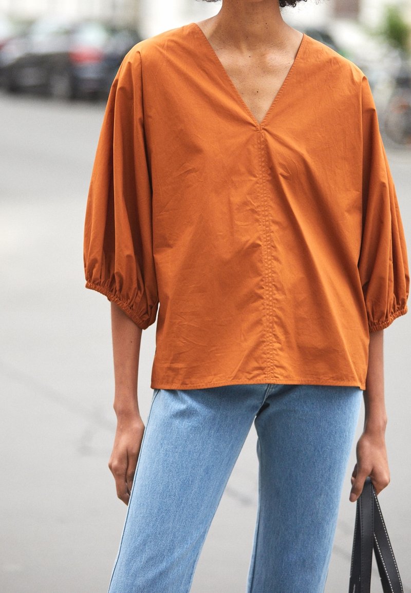Personne portant un chemisier orange rouille à col en V avec des manches bouffantes trois-quarts et un jean bleu clair, tenant un sac à main noir dans la rue.