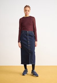 Denimrock mit einer Knopfverschluss vorne und Taschen, kombiniert mit einem langärmeligen bordeauxfarbenen Oberteil mit kleinem Muster und sportlichen Sneakers.