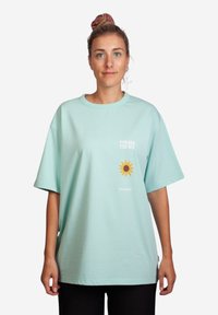 Vaalean turkoosi ylisuuri T-paita, joka on valmistettu puuvillasta; etupuolella on auringon grafiikka ja teksti "KOHTI AURINKOA", rento istuvuus, lyhyet hihat.