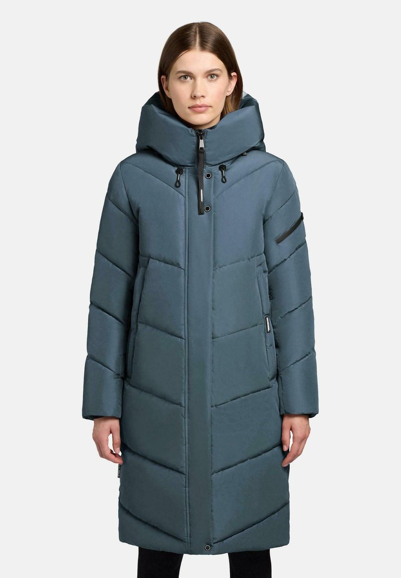 Kvinde iført lang, mørkeblå, polstret vinterjakke med stor hætte, skrå quiltet mønster, frontlynlås og lynlåslomme på venstre ærme.