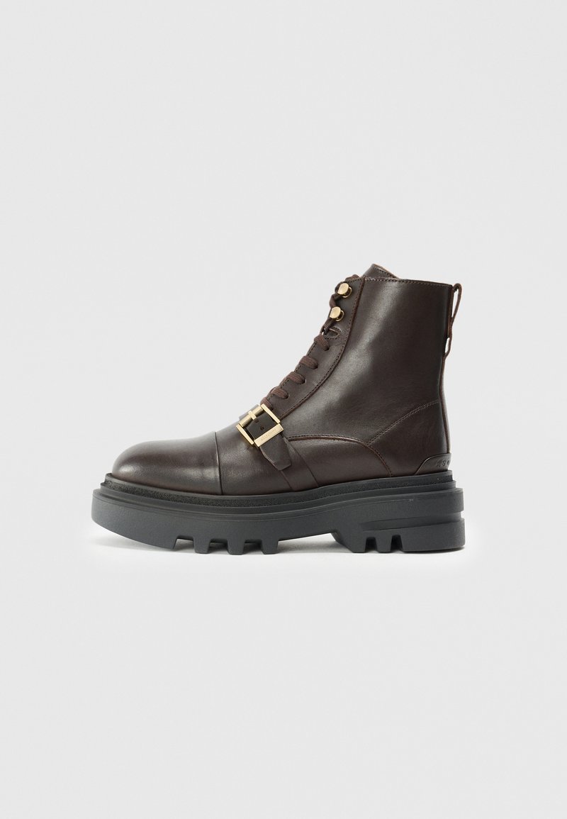 Bruine leren enkelboot met een dikke zwarte zool, vetersluiting, gouden hardware accent en een gespsluiting over de middenvoet.