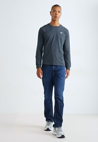 Langermet mørkegrå skjorte med rund hals, parret med blå jeans og grå joggesko, modell stående mot en ensfarget bakgrunn.