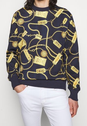 Marineblauwe sweatshirt met gouden ketting- en labelprint, gedragen met witte broek, met een ontspannen pasvorm en hand in de zak.