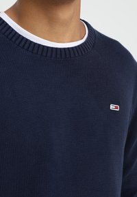 Granatowy dzianinowy sweter z prążkowanym dekoltem i białym wewnętrznym kołnierzem. Na piersi małe logo z czerwonymi i białymi akcentami.