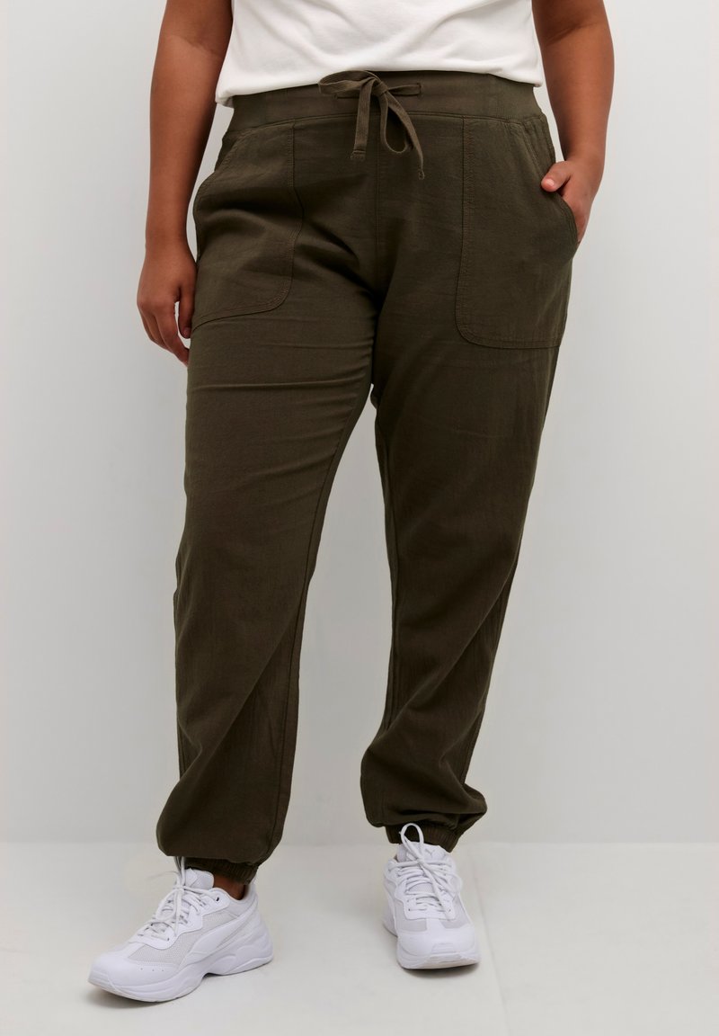 Person iført olivengrønne snorebukser med forlommer og hvide sneakers, stående mod en ensfarvet lys baggrund.