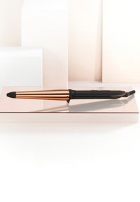 Haarkrultang met taps toelopende roségouden ton en zwart handvat, rustend op een reflecterend oppervlak tegen een lichtbeige achtergrond.