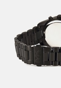 Montre noire en métal avec un bracelet en maillons, dotée d'un boîtier circulaire et d'un cadran poli. Le fermoir affiche le logo "MK".