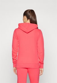 Felpa color corallo realizzata in morbido tessuto, con cappuccio a coulisse, maniche lunghe e polsini e orlo a costine. Indossata con pantaloni abbinati.