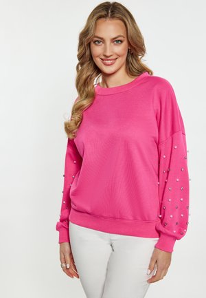 Sudadera rosa de corte holgado, con tejido texturizado, mangas en capas y cuentas decorativas a lo largo de las mangas. Llevada con pantalones blancos.