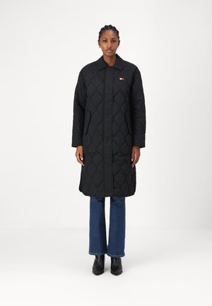 Abrigo largo acolchado negro con un cuello tipo solapa, cierre de botones a presión y un sutil detalle de logo. Combinado con jeans acampanados azules y botas negras.