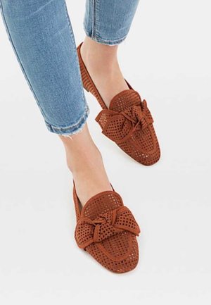 Brązowe perforowane buty typu slip-on z akcentem w postaci kokardki. Wykonane z lekkiego materiału z płaską podeszwą, w połączeniu z zniszczonymi jasno-niebieskimi dżinsami.