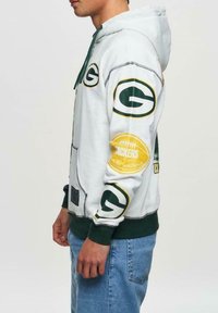 Hoodies valkoinen vihreillä yksityiskohdilla. Sisältää painettuja logoja, applikaatioita ja kengurupussin. Pehmeä, teksturoitu kangas.