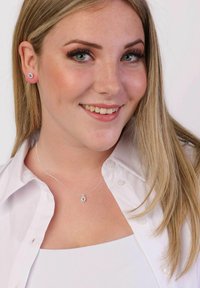 Collier en argent avec un pendentif floral présentant des accents bleus, porté avec un chemisier blanc. De petites boucles d'oreilles s'harmonisent avec le design du collier.