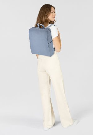 Zaino in tessuto blu con forma rettangolare, manico a contrasto e cerniere visibili. Indossato sulla schiena con pantaloni crema e calzature bianche.