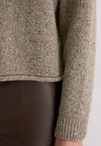 Brązowy sweter z prążkowanymi mankietami, w plamki, z prostym dołem i miękką fakturą. Noszony na ciemnobrązowej skórzanej powierzchni.
