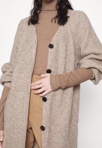 Person med en beige stickad kofta med svarta knappar över en brunspräcklig polotröja och tanbyxor, med handen vilande på midjan.