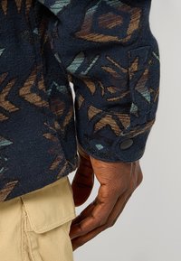 Marineblå flannel skjorte med et geometrisk mønster i brune og teal nuancer, har en knappet manchet detalje og en robust stoftekstur.